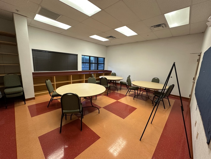 Multi-purpose classroom with long tables, chairs, and supplies arranged along the windows